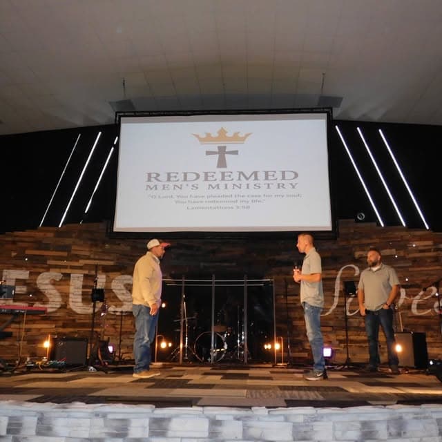 Three men stand on a stage before a screen displaying the Redeemed Men's Ministry logo.